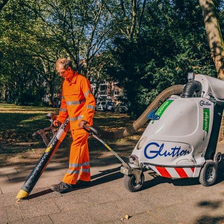 Mann im orangenen Arbeitsanzug bedient Abfallsauggerät auf Plastersteinen.