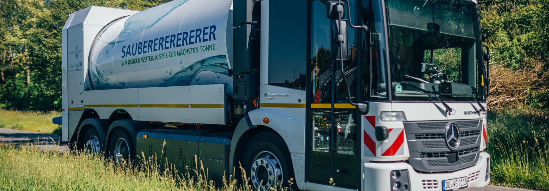 Weißes Müllfahrzeug auf Straße, vorne auf dem Bild Wiese, hinten Bäume und blauer Himmel