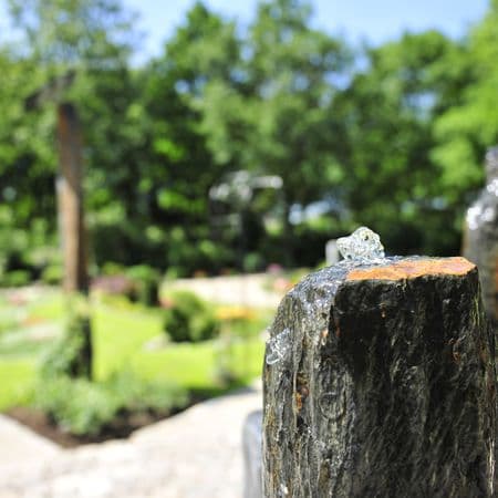 Memoriam Garten auf dem Friedhof, Gräber im Hintergrund, eine Brunenstele im Vordergrund.