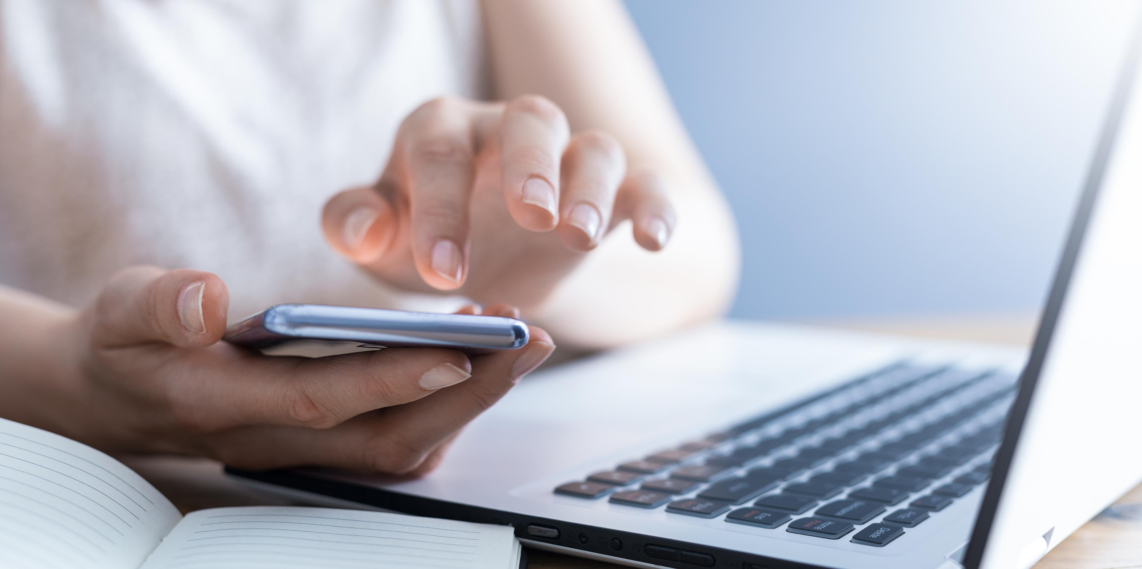 Frau hält Smartphone in der Hand, davor liegen ein Notebook und ein Heft