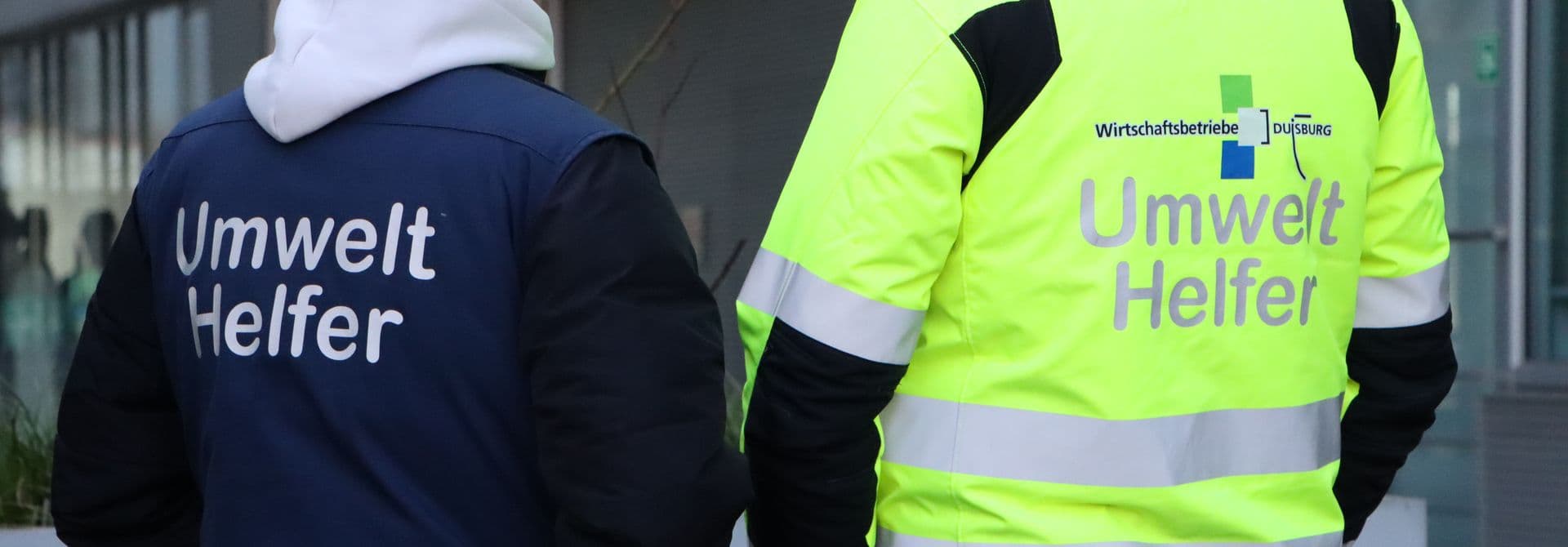 Ein Mitarbeiter in einer blauen und ein Mitarbeiter in einer gelben Jacke mit der Aufschrift Umwelthelfer