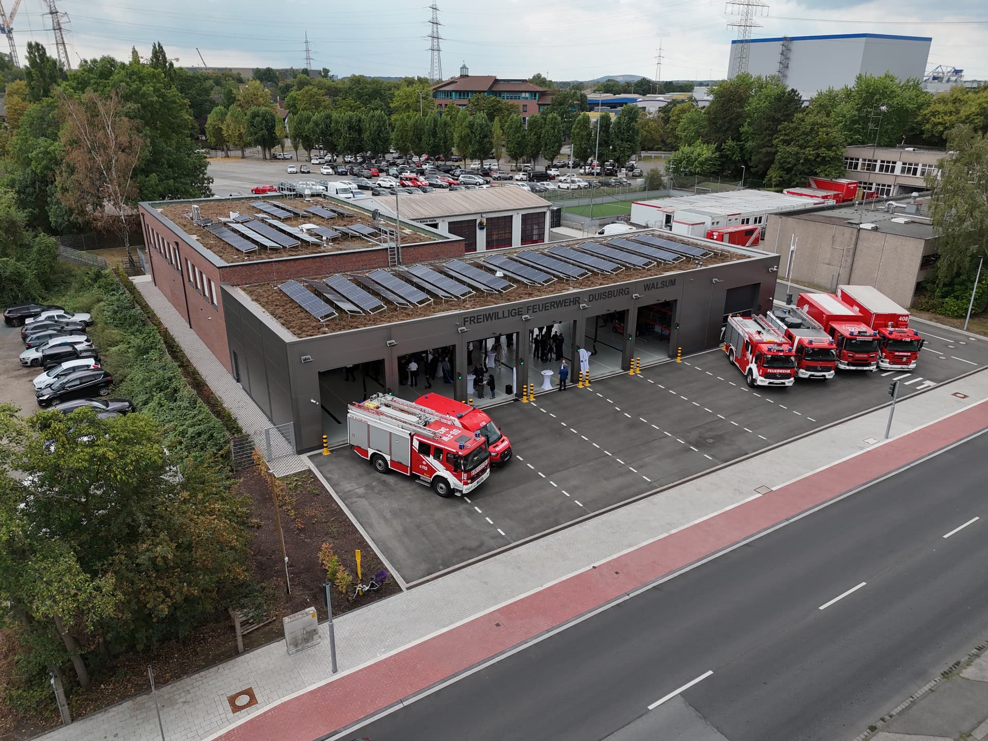 Feuerwache mit roten Feuerwehrfahrzeugen, grüner Dachbegrünung und Solarpaneelen, umgeben von Bäumen und Parkflächen.