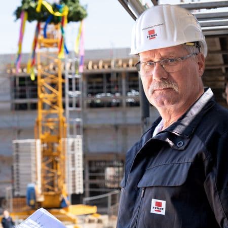 Ein Mann mit Helm steht auf einer Baustelle; im Hintergrund ein Kran mit Richtkranz.