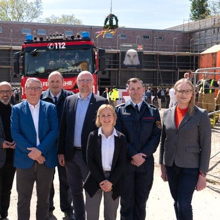 Eine Gruppe von Menschen steht vor einem Feuerwehrwagen auf einer Baustelle mit einem Richtkranz