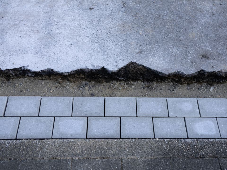 Man sieht von oben eine Abbruchkante aus grauem Asphalt und graue gepflasterte Steine.