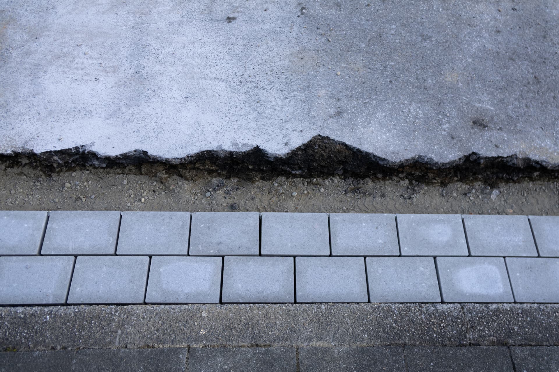Man sieht von oben eine Abbruchkante aus grauem Asphalt und graue gepflasterte Steine.