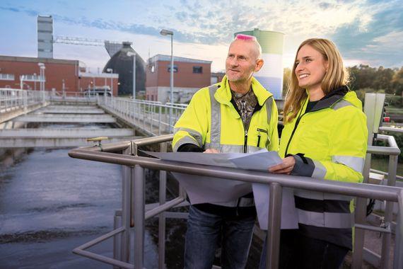 Motiv Kläranlage Eine Mitarbeiterin und ein Mitarbeiter in gelber Arbeitsjacke schauen über eine Kläranlage und halten einen Plan in den Händen