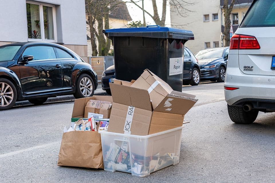 Altpapiersammlung Eine graue Mülltonne mit blauem Deckel ist zu sehen, davor stapelt sich Pappe und Papier.