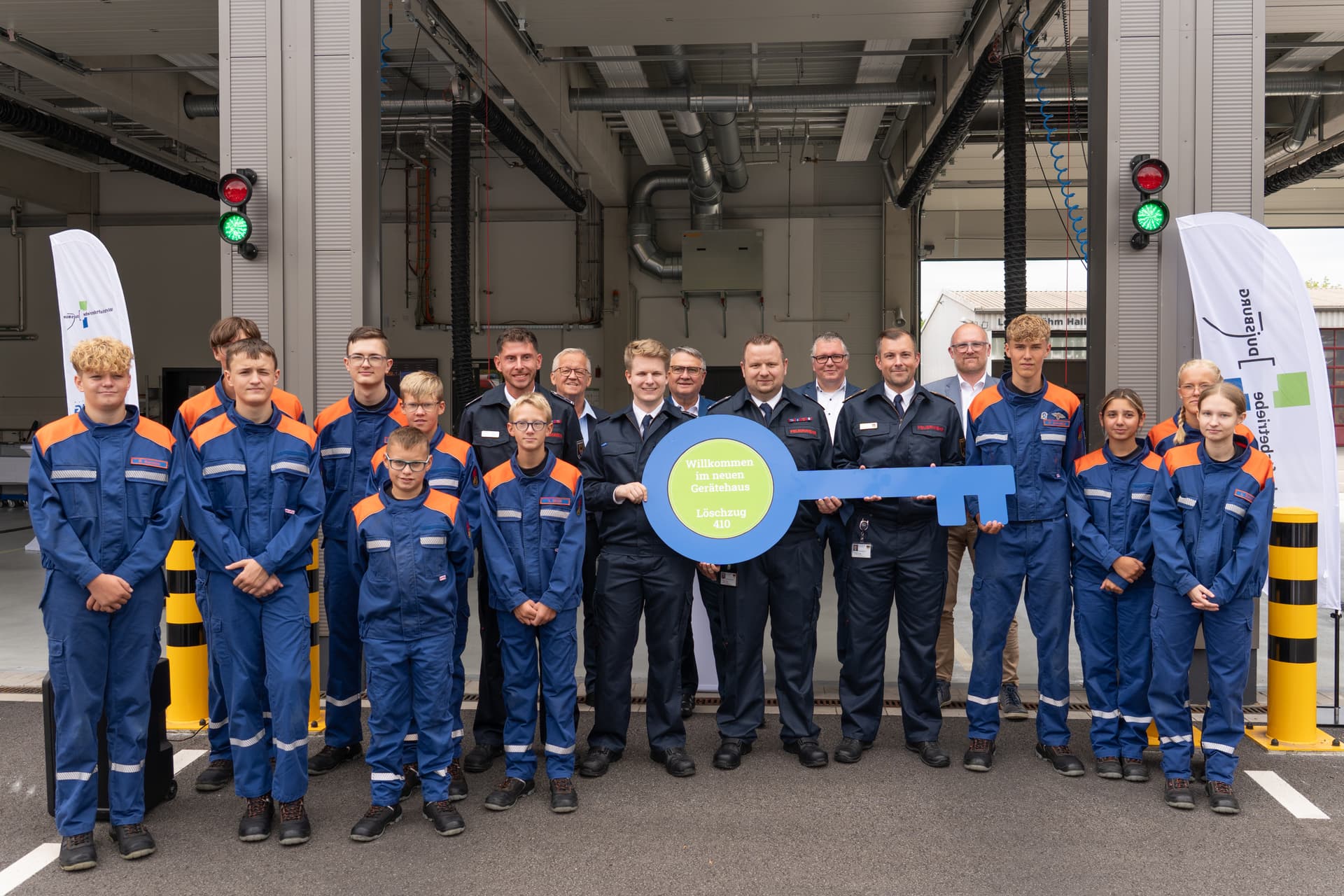 Gruppe von Feuerwehrleuten in blauen Uniformen hält einen großen Symbolschlüssel vor einem neuen Gerätehaus.