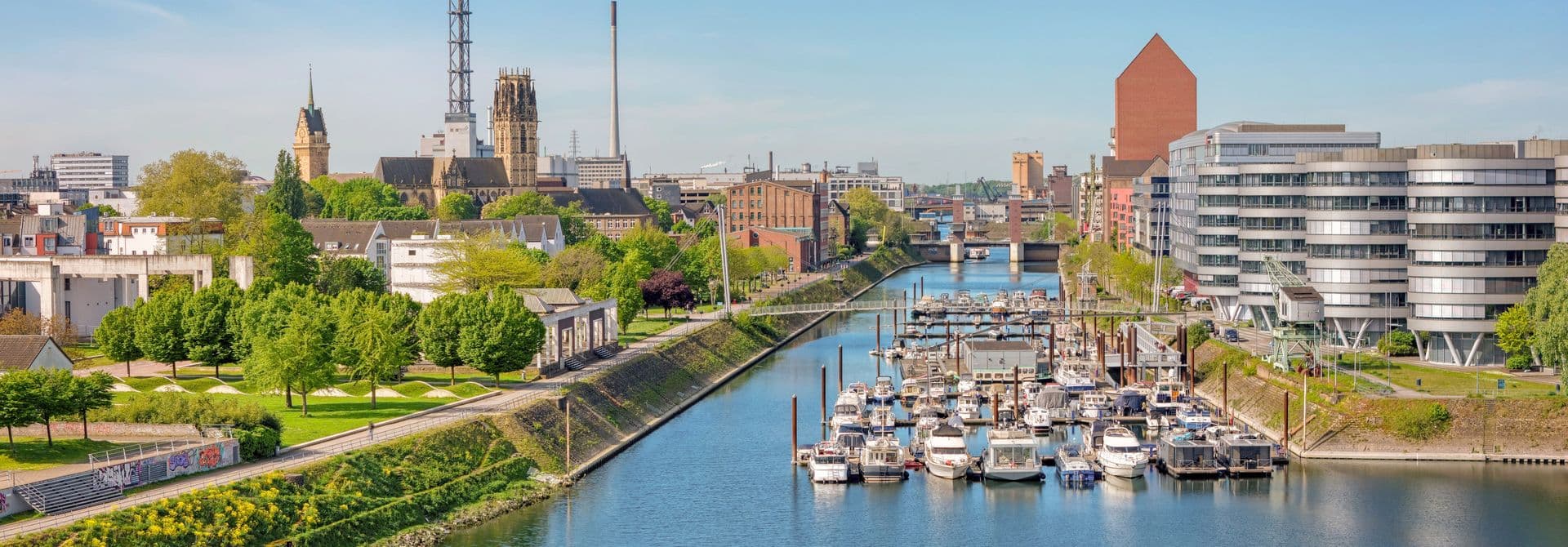 Stadtpanorama Duisburger Stadtansicht mit Fluss, Hafen mit Booten, modernen Gebäuden rechts und historischen Bauten links, blauer Himmel im Hintergrund.