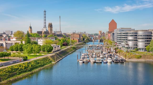 Duisburger Stadtansicht mit Fluss, Hafen mit Booten, modernen Gebäuden rechts und historischen Bauten links, blauer Himmel im Hintergrund.